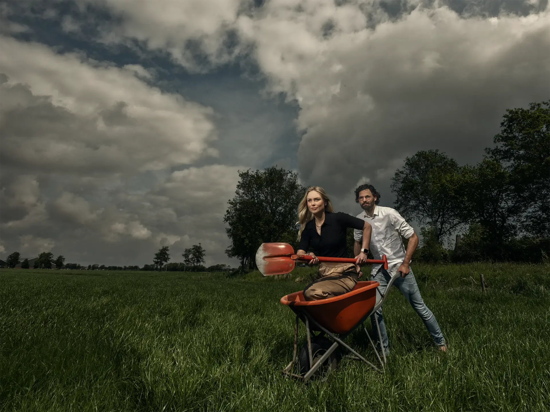 Man in weiland met vrouw in kruiwagen net schep in handen